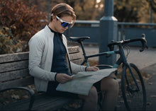 A person sitting on a wooden bench outdoors, reading a map, with a bicycle beside them. The person is wearing white-framed sunglasses with blue lenses, a light gray jacket, and a dark top.