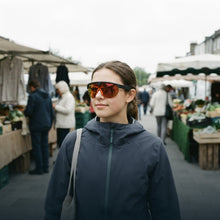 A woman wearing reflective orange-tinted shield sunglasses and a dark navy zip-up jacket walks through an outdoor farmers market with stalls and shoppers blurred in the background.