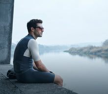 A man wearing a dark gray and white short-sleeve athletic wetsuit sits on a concrete ledge next to a calm river, looking out towards the water under a misty, overcast sky.