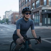 A woman wearing a black bicycle helmet, sunglasses, and a dark gray short-sleeve cycling jersey rides a road bike on an urban city street with brick buildings in the background.
