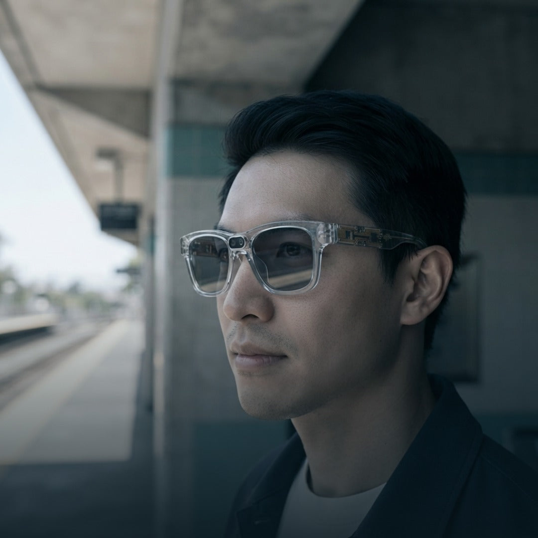 A man in profile wearing clear-framed smart sunglasses with dark tinted lenses, standing at a train station platform.