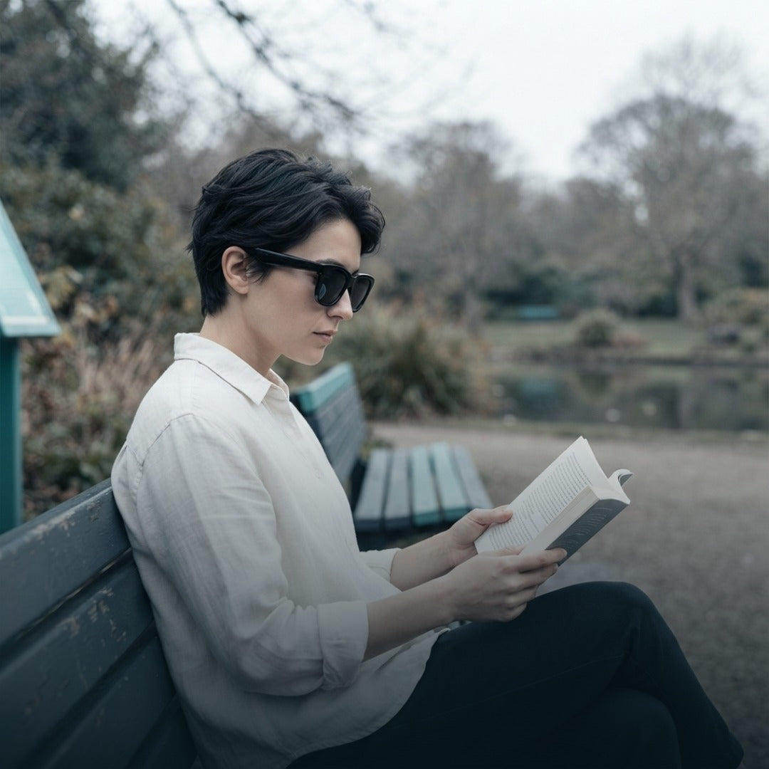 A person with short dark hair wearing black smart sunglasses, a white button-up shirt, and black pants sits on a park bench reading a book. The background shows a blurred outdoor park scene with trees and a pond.
